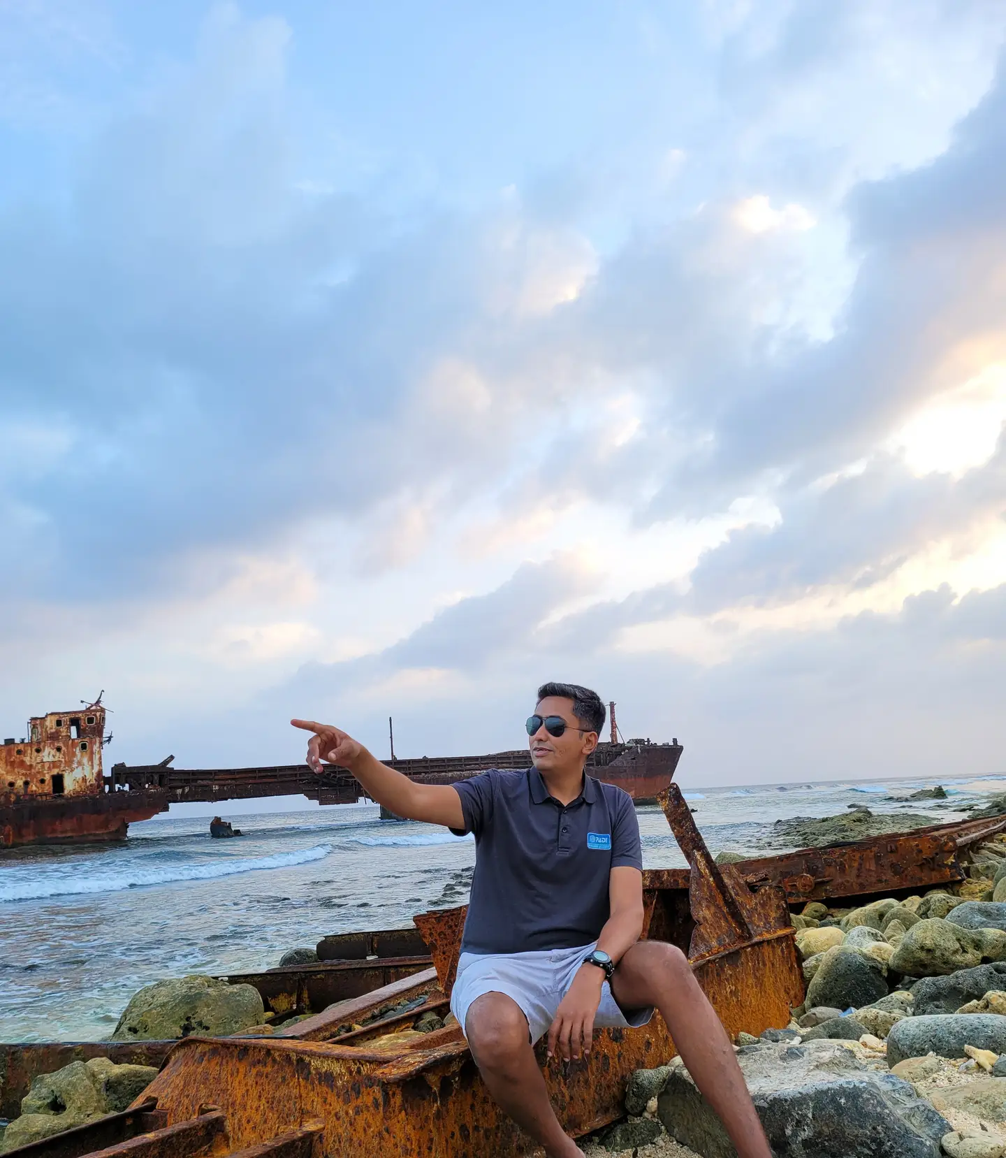 Hardik Datanwala seated on rusted wreckage on a rocky shore, pointing towards a beached shipwreck lying offshore under a wide evening sky